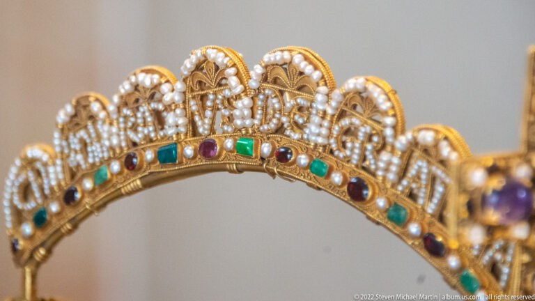 Close view of a gold crown band with scalloped openwork arches, rows of pearls, and green and purple stones.