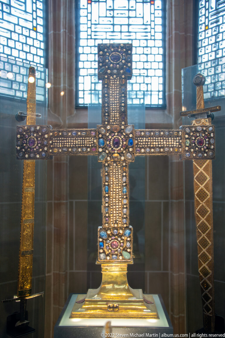Large upright gold processional cross covered with pearls and colored gemstones in a museum case.
