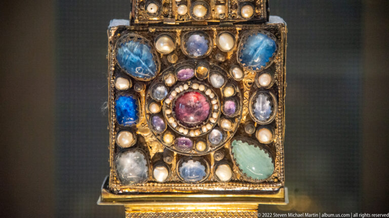 Rectangular gold treasury detail with a central red stone, pearl ring, and large blue, green, and pale gemstones.