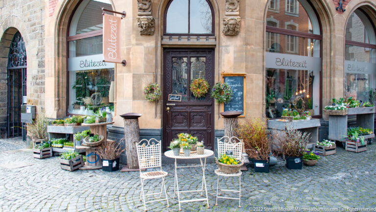 Flower shop with potted plants, crates, and white chairs arranged outside a carved doorway on a cobbled street.