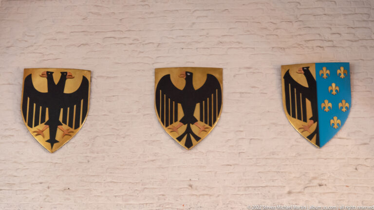 Three wall-mounted heraldic shields, two with black eagles and one combining an eagle with gold fleurs-de-lis on blue.