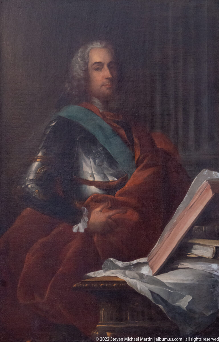 Dark portrait of a seated man wearing a blue-green sash beside papers and a large open book.