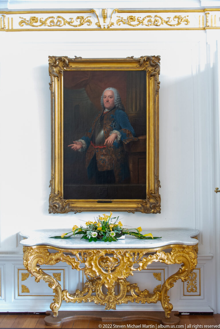 Framed portrait of a seated man above an elaborate gilded console table with floral decoration.