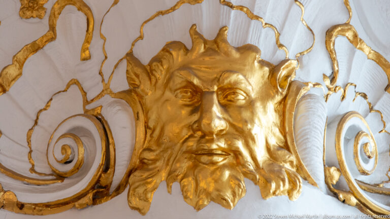 Close view of a gilded bearded face set within curling white-and-gold stucco ornament.