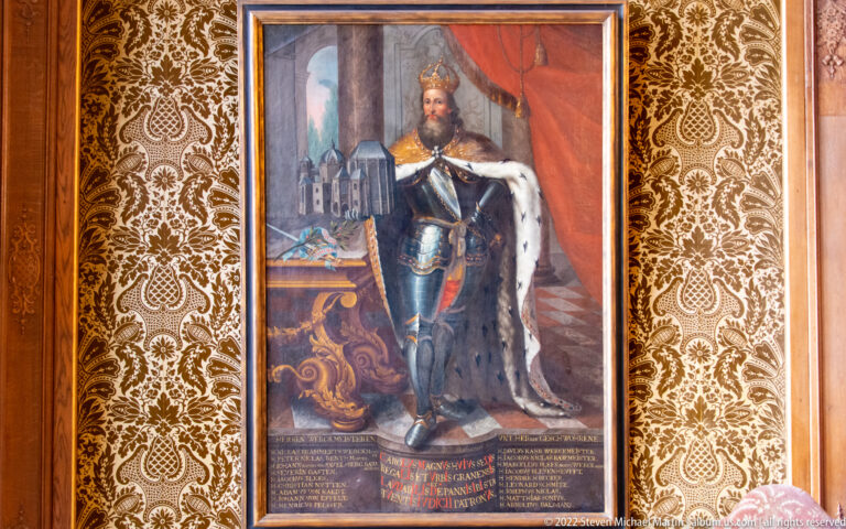 Full-length painted ruler in armor, crown, and ermine-lined mantle beside a model of a large building.
