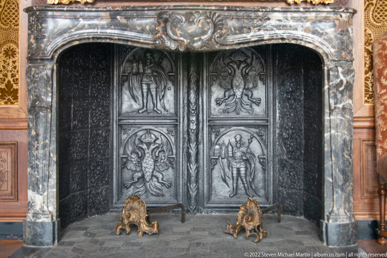 Large dark fireplace with relief panels, marble surround, heraldic carving, and two gilded fire supports.