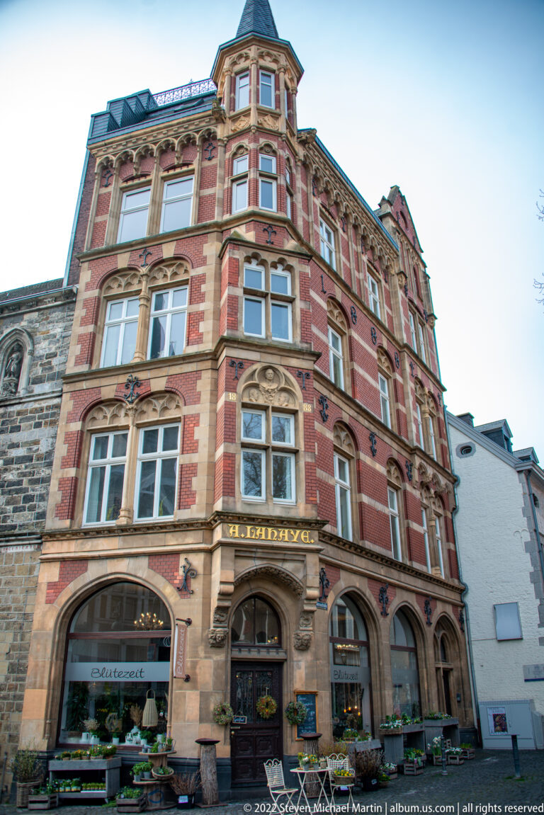 Ornate red-and-stone corner building with arched storefronts, bay windows, carved trim, and café seating.