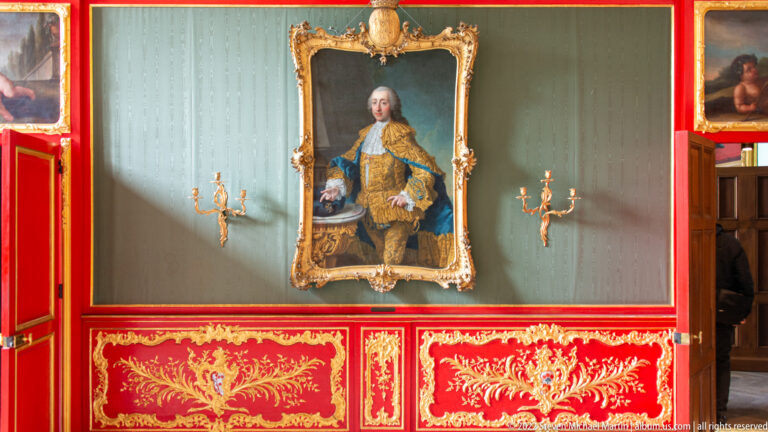 Formal portrait of a man in an embroidered coat, displayed between gilt sconces above red-and-gold paneling.