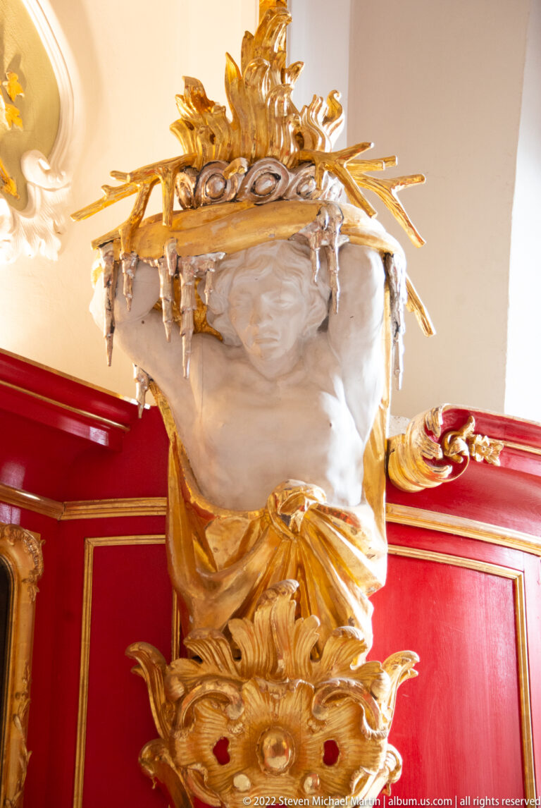 White male architectural figure straining beneath a broad gilded burden above a red-and-gold wall.