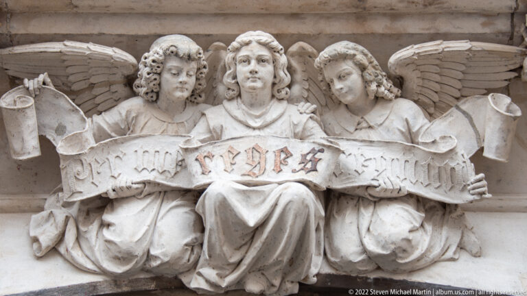 Three carved angels holding a long inscribed scroll, with wings spread behind them.