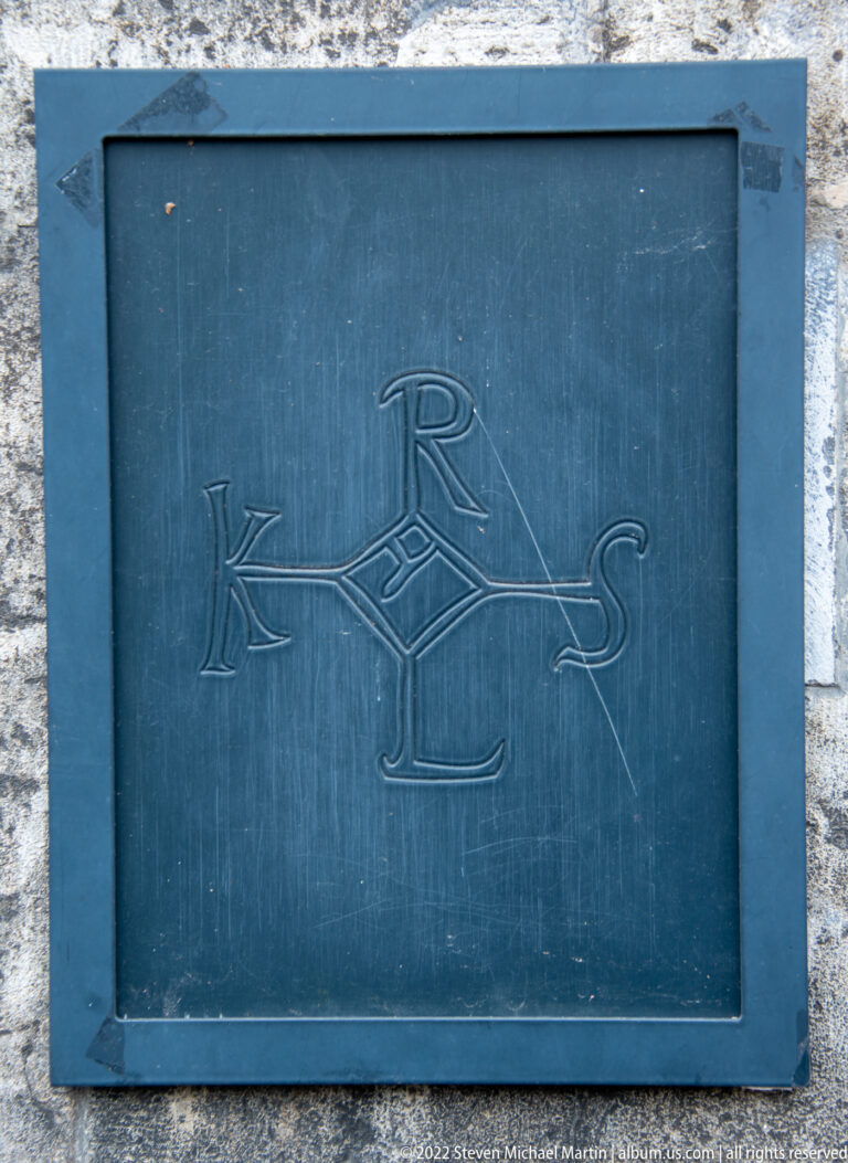 Dark rectangular metal plaque engraved with an interlaced monogram of several Roman letters.