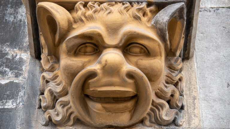 Close view of a carved stone grotesque with pointed ears, curling hair, heavy brows, and a broad grin.