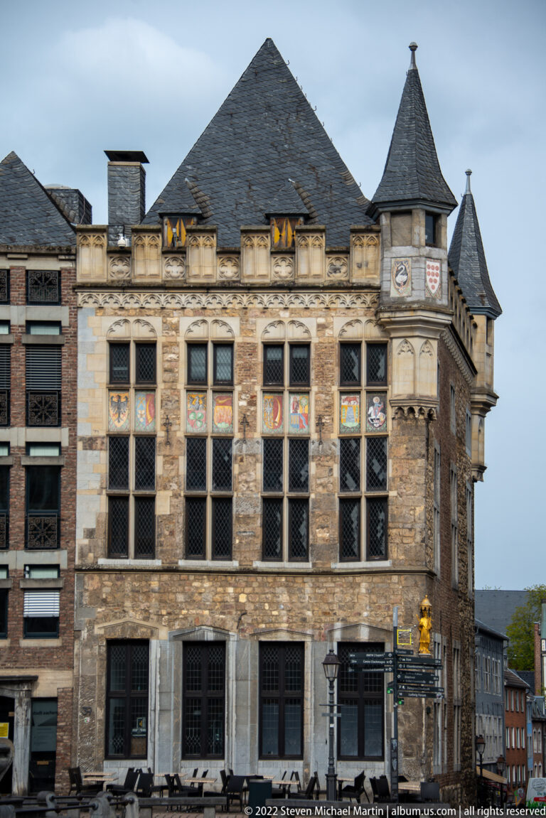 Stone civic building with tall mullioned windows, steep roofs, corner turrets, and colorful heraldic shields.
