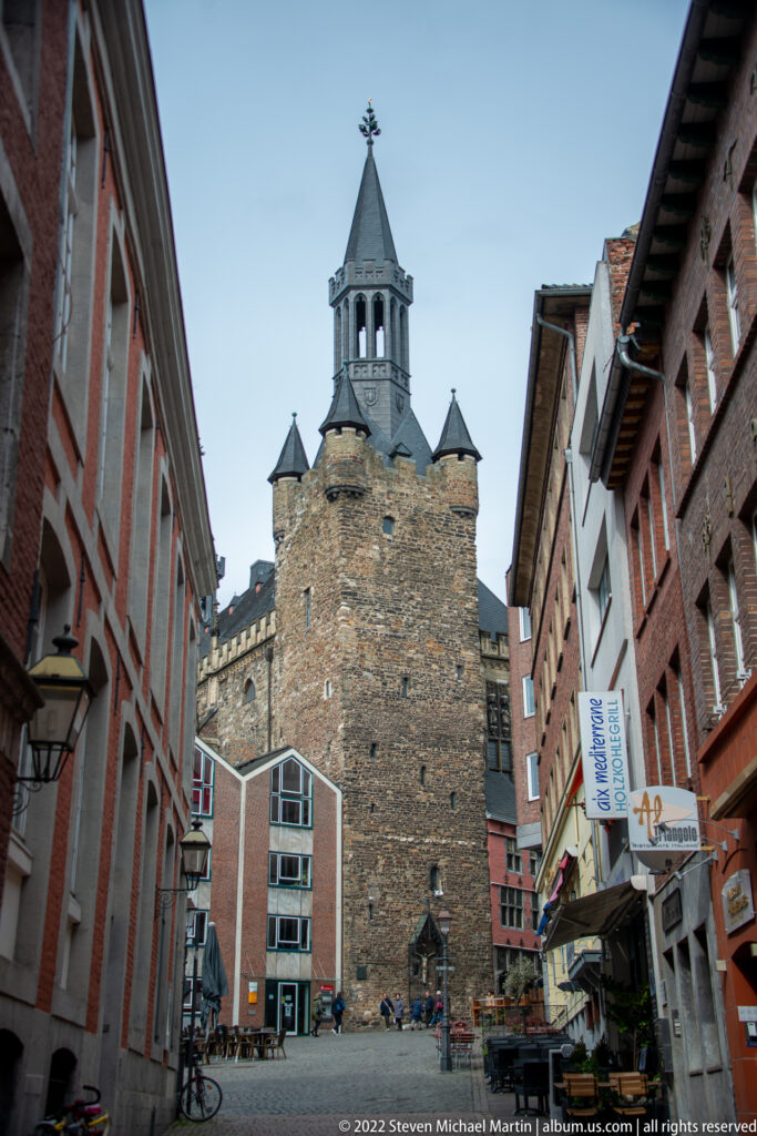Massive medieval stone gate tower with steep roof and small turrets at the end of a narrow Aachen street.