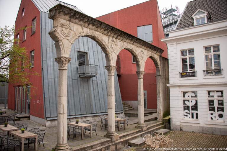 Freestanding row of carved stone arches on slender columns in a modern courtyard.