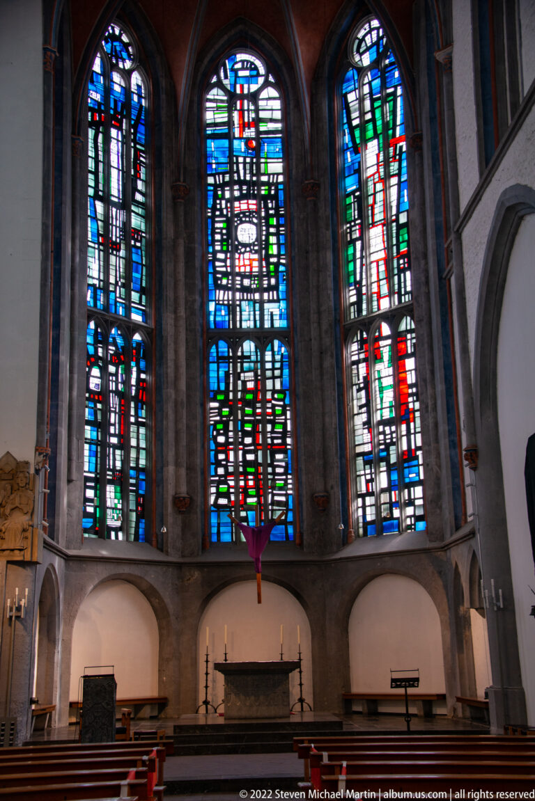 Three tall stained-glass windows above a simple altar and shallow arched recesses inside a church.