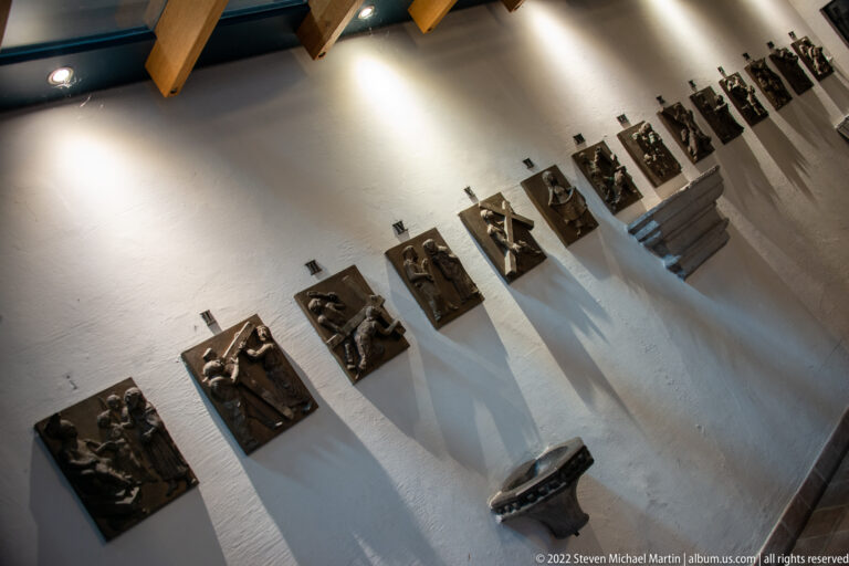 Row of small bronze religious relief panels mounted sequentially along a white church wall.