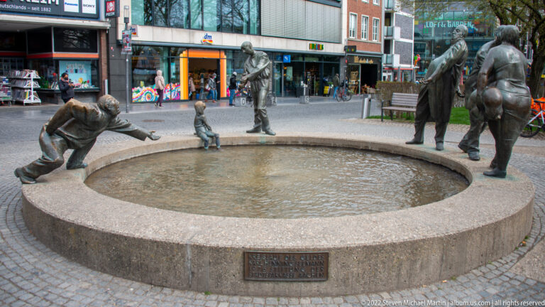 Circular stone fountain surrounded by playful bronze figures of adults and children in a pedestrian shopping area.