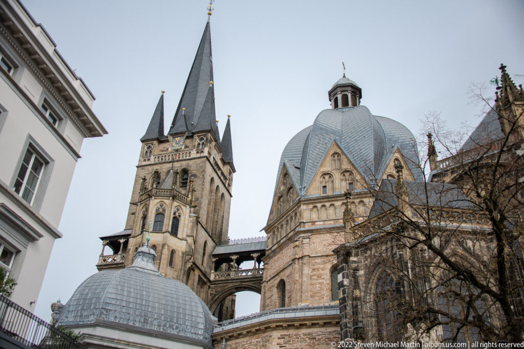 Close exterior view of Aachen Cathedral with tall tower, octagonal dome, chapels, and layered historic roofs.
