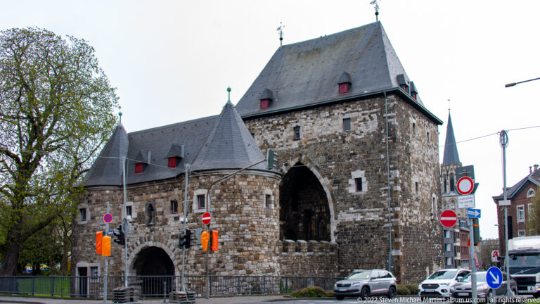Large medieval stone gatehouse with pointed passage, steep roof, and modern traffic signs in Aachen.
