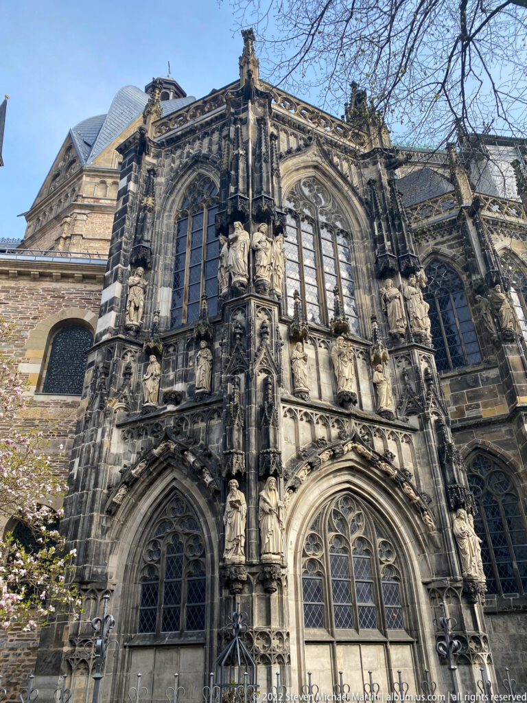 Low-angle exterior view of Aachen Cathedral's Gothic choir with tall windows, buttresses, pinnacles, and saint statues.