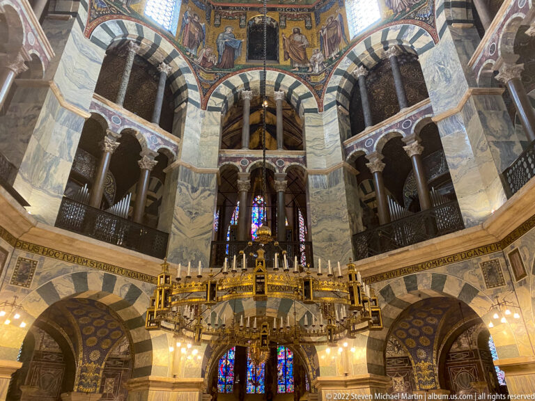 Interior of Aachen Cathedral's Octagon with striped arches, upper galleries, mosaics, and the circular Barbarossa Chandelier.