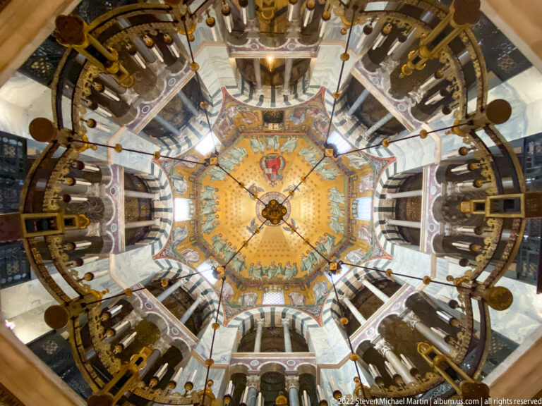 Wide upward view of Aachen Cathedral's Octagon with golden cupola, galleries, columns, arches, and circular chandelier.