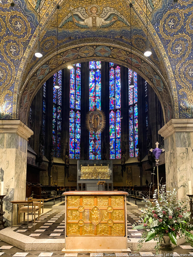 Altar before tall stained-glass windows, framed by a richly decorated arch and marble piers inside Aachen Cathedral.
