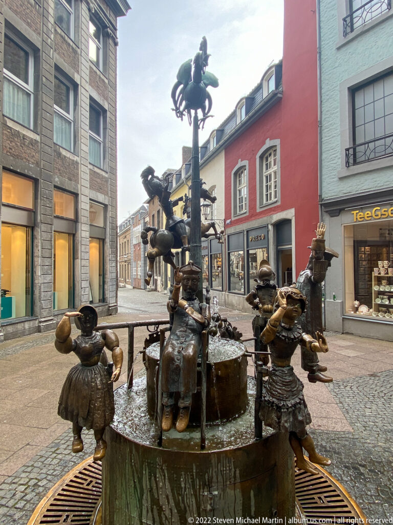 Bronze figures of the Puppenbrunnen grouped around a circular fountain in an Aachen pedestrian street.
