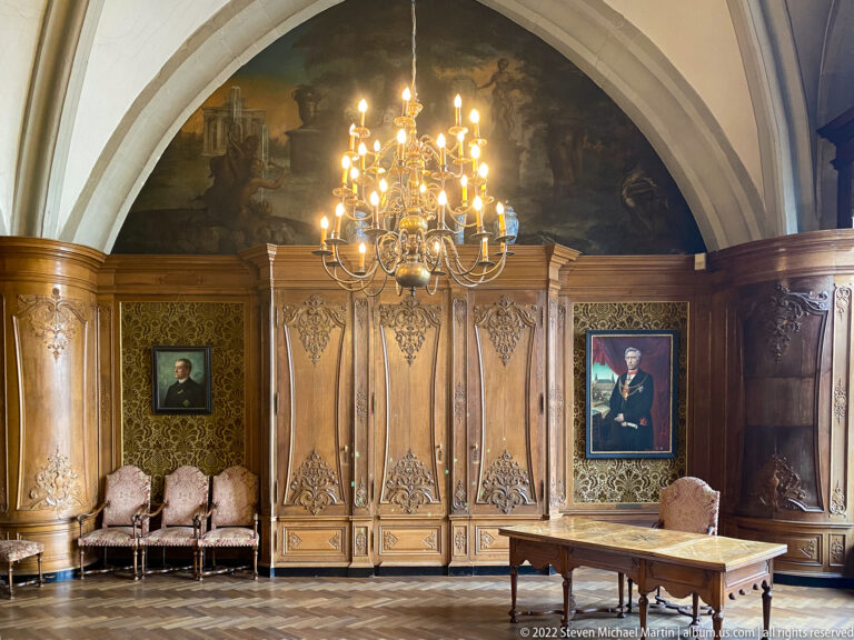 Wood-paneled historic room with crystal chandelier, painted vault, formal portraits, chairs, and a writing table.