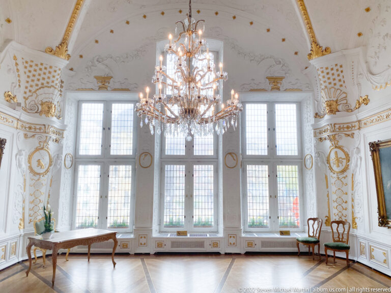 White-and-gold Rococo room with crystal chandelier, tall windows, parquet floor, and light period furniture.