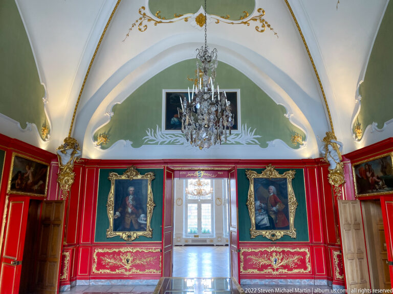 Red-and-green historic room with gilded paneling, framed portraits, crystal chandelier, and doorway to a brighter adjoining room.