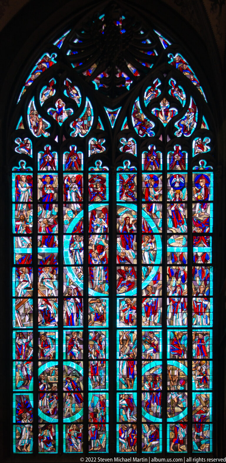 Large multi-light stained-glass window glowing in red, blue, turquoise, and white within a dark cathedral interior.