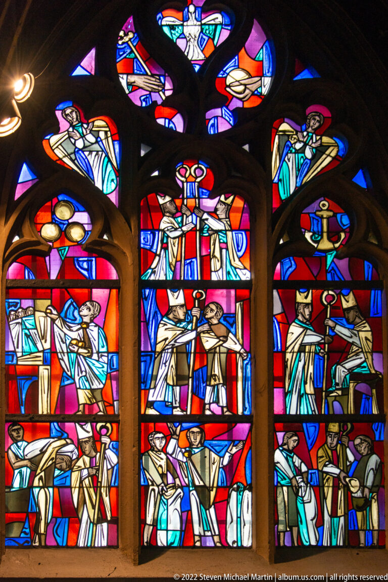 Three-part stained-glass window filled with multiple sacred scenes and smaller figurative tracery panels above.