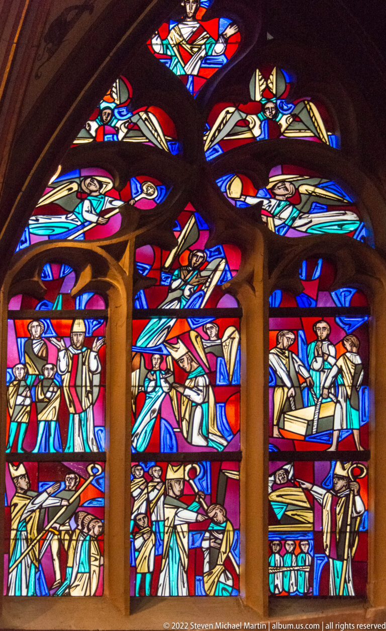 Three tall stained-glass panels crowded with biblical figures beneath smaller colored tracery panels.