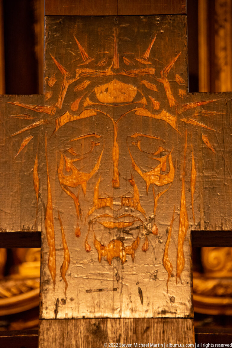 Incised face of Christ with a crown of thorns carved into a weathered wooden cross beam.