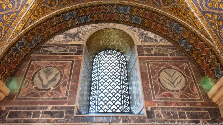 Arched lattice window framed by colored marble panels and a patterned mosaic border inside Aachen Cathedral.