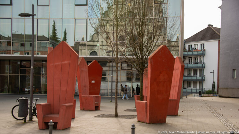 Red faceted sculptural street furniture stands in a paved plaza beside modern glass-fronted buildings and trees.