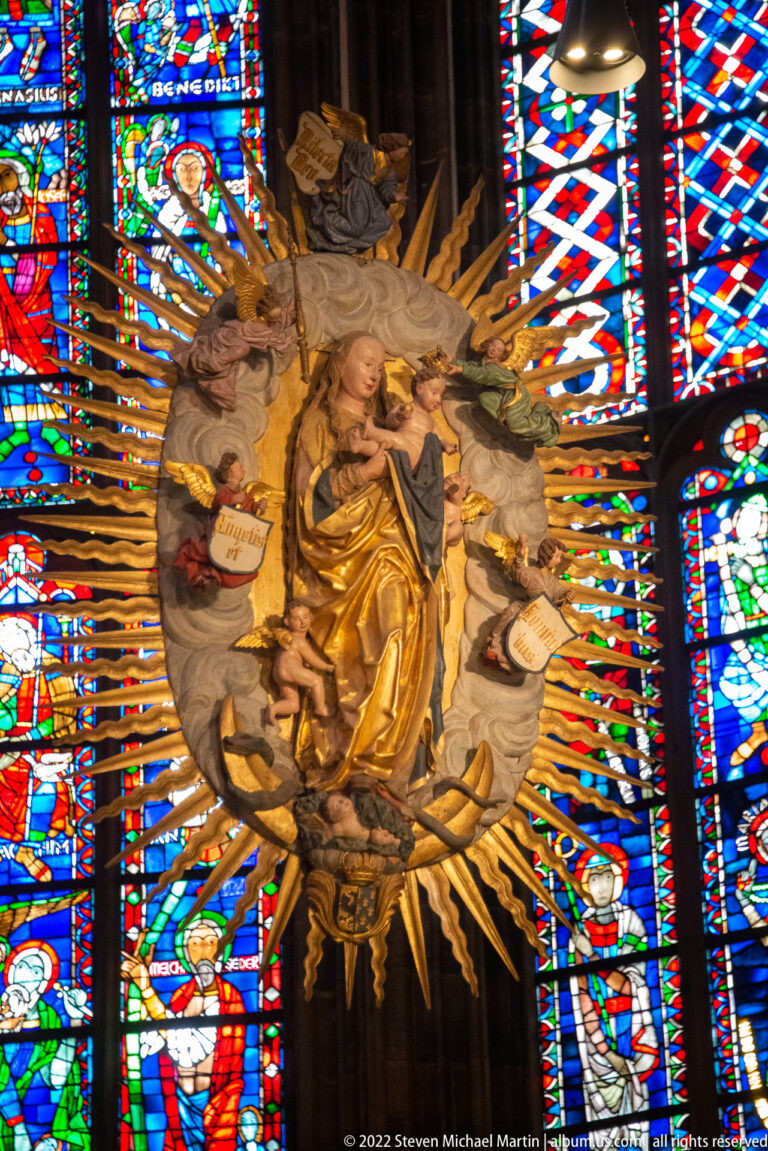 Gilded Virgin and Child with angels and radiating metal rays set before colorful stained-glass windows.