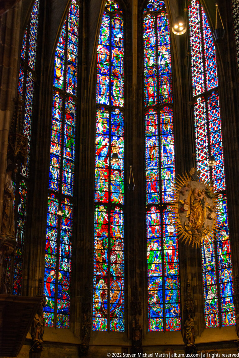 Tall stained-glass windows between dark stone piers, with a gilded radiant devotional object at lower right.