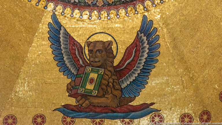 Winged lion with halo and colorful wings holding a decorated book against a gold mosaic background.