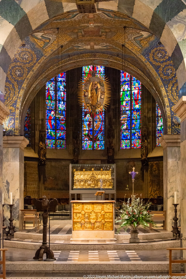 Cathedral sanctuary under a decorated stone arch, with gilded altar furnishings, candles, flowers, and stained glass beyond.