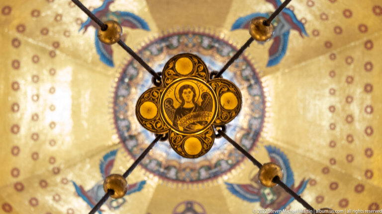 Gilded circular medallion suspended on dark rods beneath a gold mosaic ceiling.