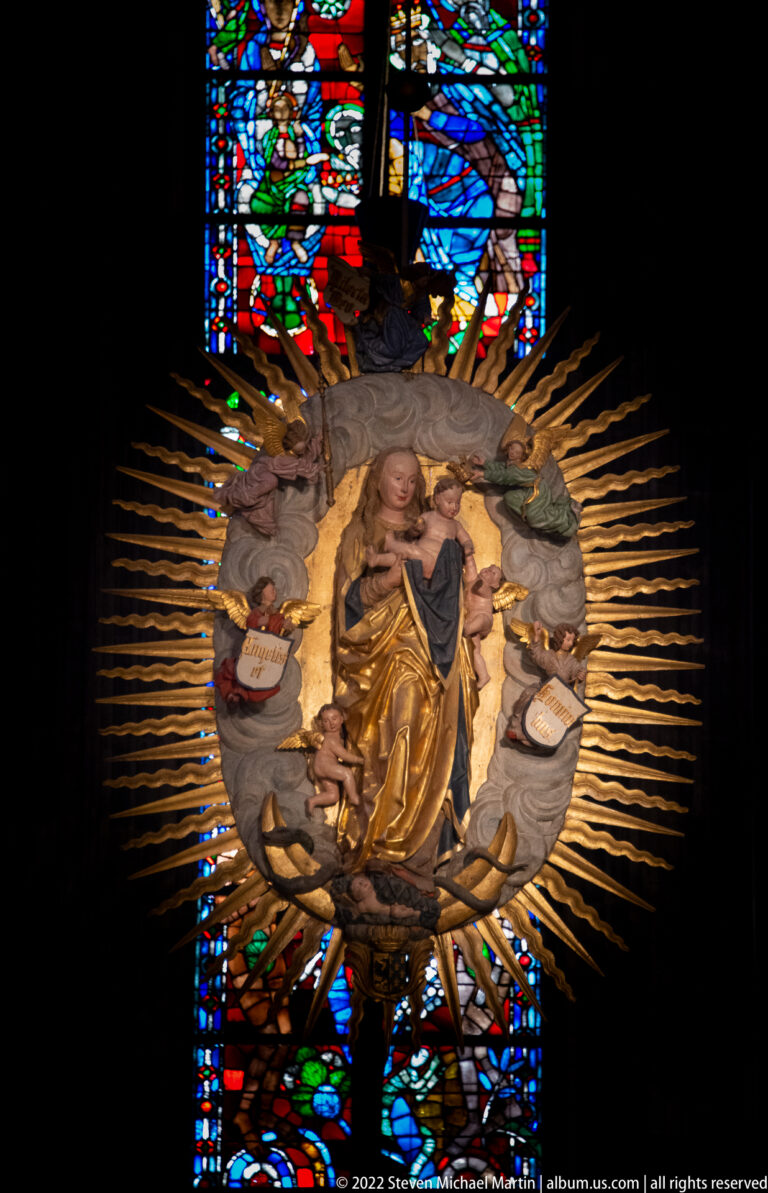 Radiant gilded Virgin and Child with angels suspended against dark stained-glass windows.