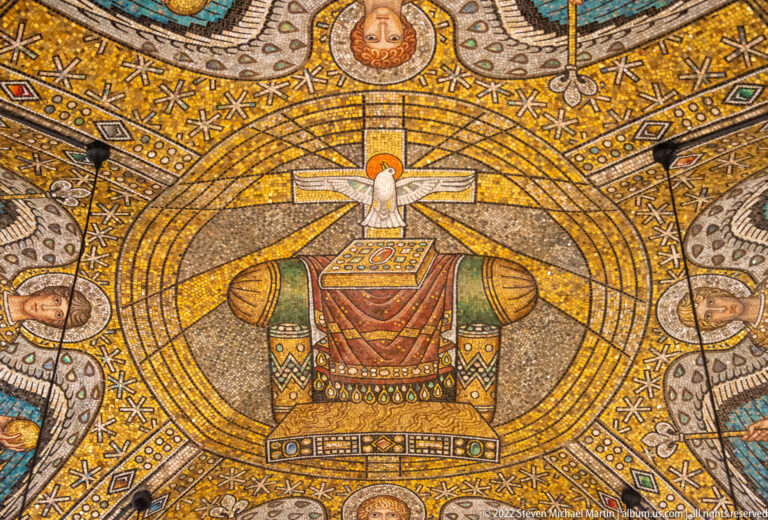 Close mosaic view of a patterned throne-like seat beneath a white winged figure on a gold ground.