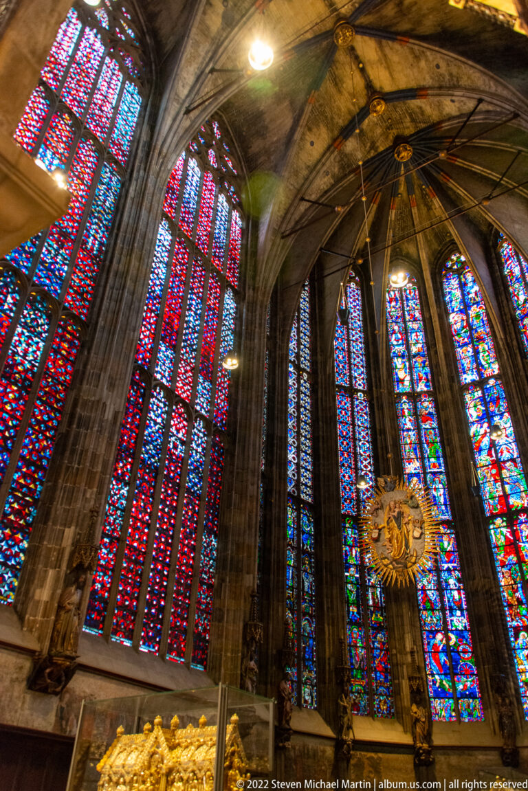 Low-angle view of tall stained-glass choir windows beneath radiating stone vault ribs, with gilded ornament below.