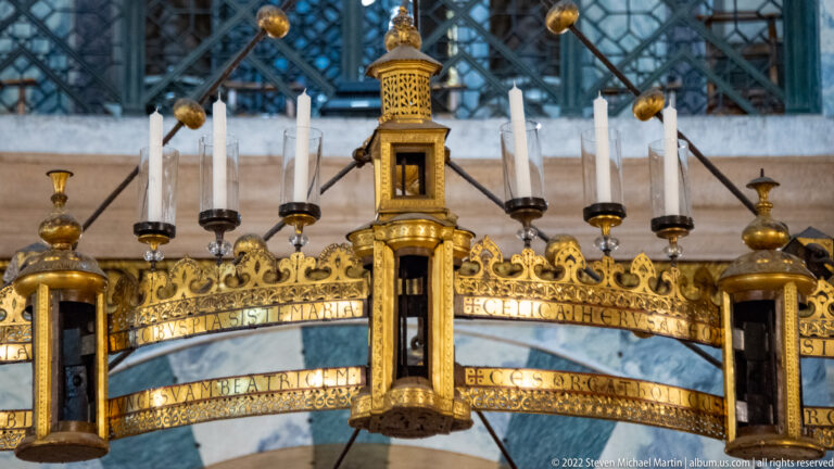 Close view of a gilded ring chandelier with white candles, tower-like fixtures, inscriptions, and dark suspension rods.
