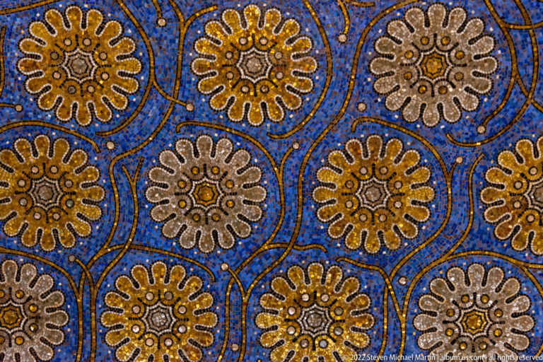 Repeating white and gold rosettes linked by curving gold lines on a deep blue mosaic ground.