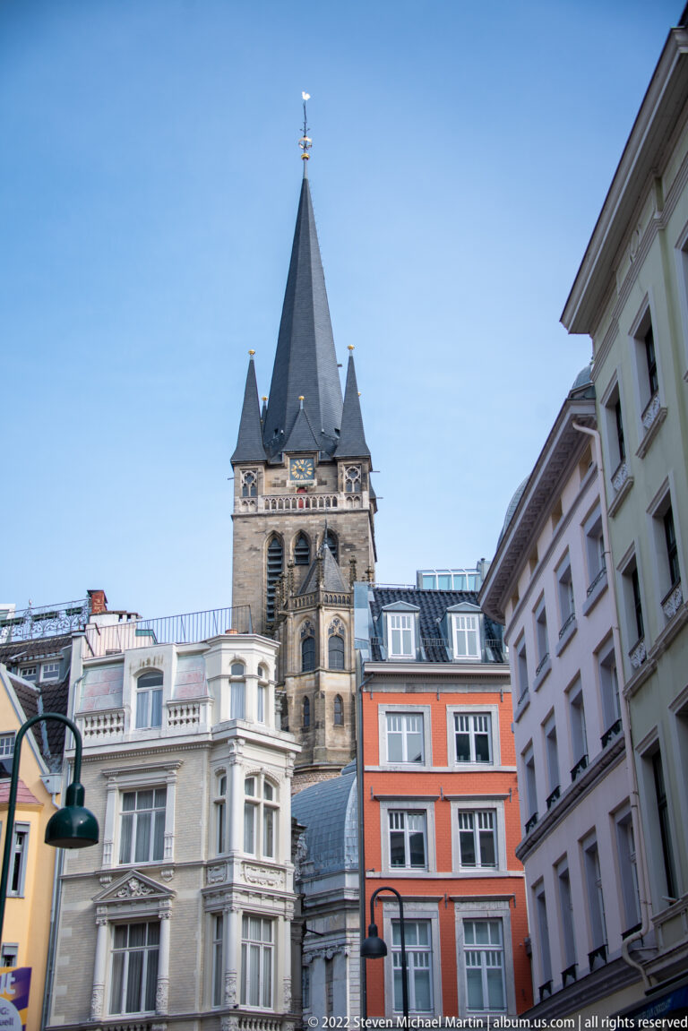 Gothic church tower with a steep spire and corner pinnacles rising above colorful town buildings.