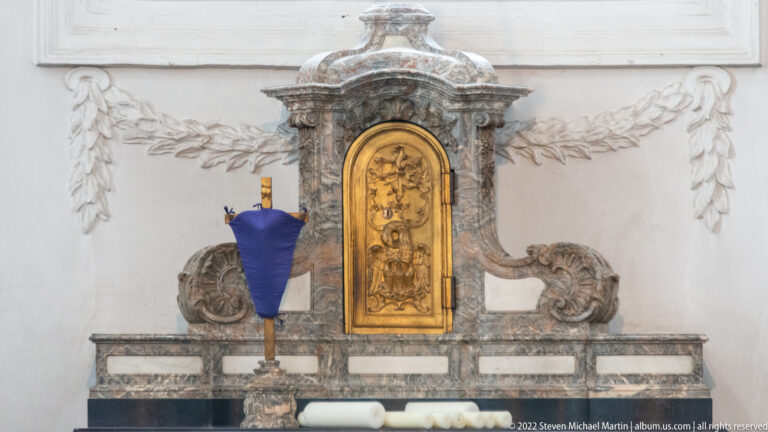 Ornate gray-marble wall furnishing with central gilded door or plaque and sculpted supports on both sides.